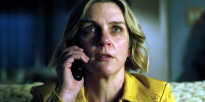 Rhea Seehorn, wearing a yellow shirt, is on the phone in Vince Gilligan's Pluribus. Her eyes are glazed as if she's been crying or is very tired.