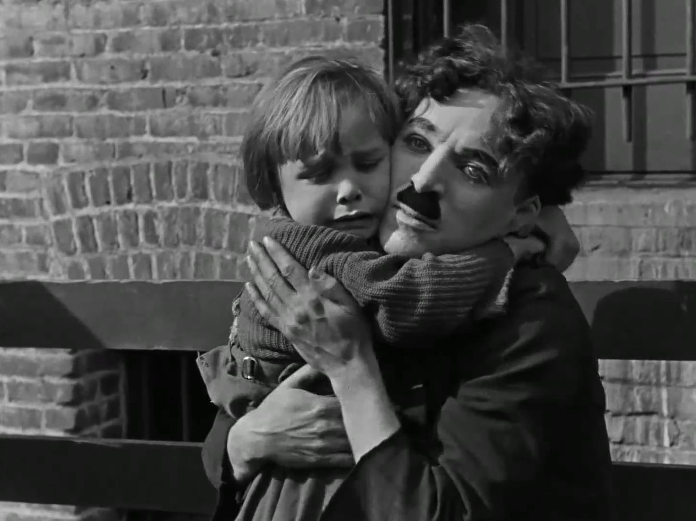 Charlie Chaplin, sporting his famous moustache, carries a small child, appearing to comfort the upset child, in the silent, black and white film, The Kid.
