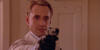 Roy Scheider points a pistol at someone off screen while wearing a black glove in the film, Cohen & Tate.