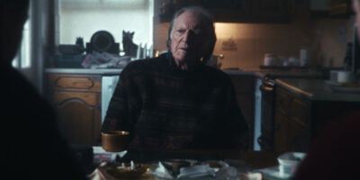 A somber, cinematic mid-shot of actor David Bradley in the film 'Brothers.' He looks off-camera with an expression of intense grief and moral conflict, illuminated by soft light against a dark, blurred background.