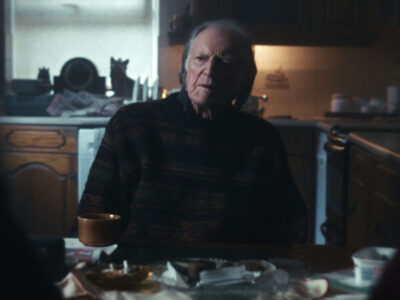 A somber, cinematic mid-shot of actor David Bradley in the film 'Brothers.' He looks off-camera with an expression of intense grief and moral conflict, illuminated by soft light against a dark, blurred background.