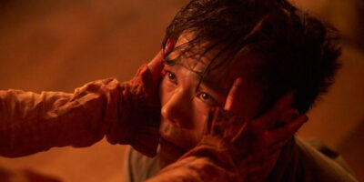 A man, lit in a red-orange fiery glow, looks in pain and fear as two hands grip his temples.