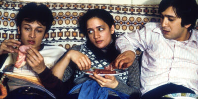 Jimi Mistry as Tariq "Tony" Khan in the 1999 film, East is East, sits on a 1970s couch alongside fellow actors Chris Bisson and Archie Panjabi.