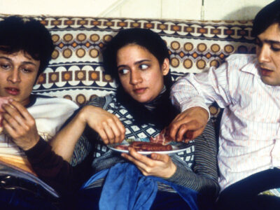 Jimi Mistry as Tariq "Tony" Khan in the 1999 film, East is East, sits on a 1970s couch alongside fellow actors Chris Bisson and Archie Panjabi.