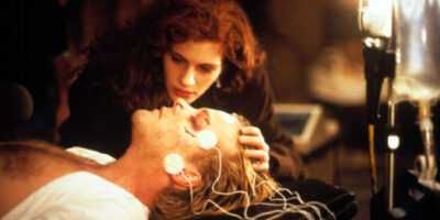 Julia Roberts nurses an unconscious Kiefer Sutherland, who lies on a hospital bed with monitors attached to his head and face, in the film, Flatliners.