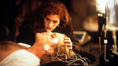 Julia Roberts nurses an unconscious Kiefer Sutherland, who lies on a hospital bed with monitors attached to his head and face, in the film, Flatliners.