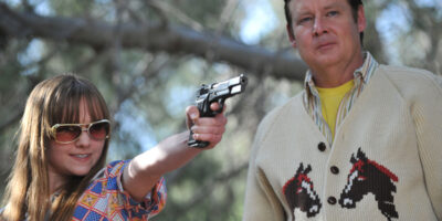 A young woman, wearing sunglasses and pointing a handgun towards the camera, stands next to an older man in the film, God Bless America.
