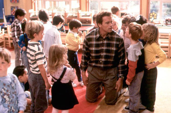 Arnold Schwarzenegger, sitting on his knees on a primary school carpet, is surrounded by children in the film, Kindergarten Cop.