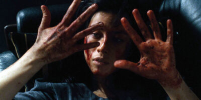 A bloodied young woman stares at the camera with her hands held up, her fingers showing signs of torture.