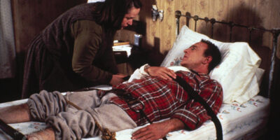 Kathy Bates as Annie Wilkes in the film, Misery, adjusts the straps she's using to tie Paul Sheldon (James Caan) to a bed.
