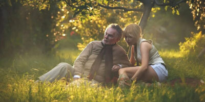 Ellie Bamber and Derek Jacobi sit on the ground in a summer garden surrounded by yellow flowers in the film, Moss and Freud.