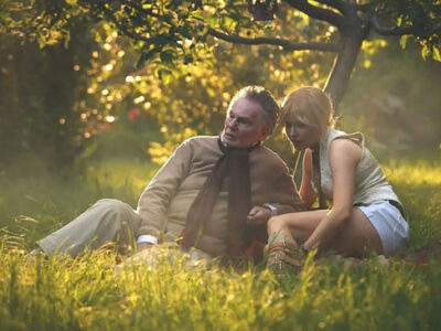 Ellie Bamber and Derek Jacobi sit on the ground in a summer garden surrounded by yellow flowers in the film, Moss and Freud.