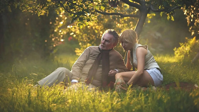 Ellie Bamber and Derek Jacobi sit on the ground in a summer garden surrounded by yellow flowers in the film, Moss and Freud.