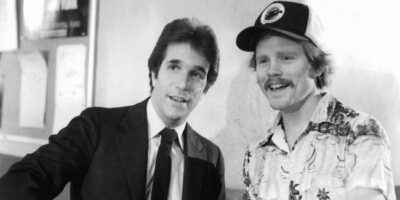 A black and white image depicting Henry Winkler in a suit and in-character during the making of the 1982 film Night Shift talking with director Ron Howard who is wearing a casual shirt and baseball cap.