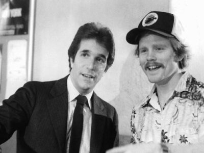 A black and white image depicting Henry Winkler in a suit and in-character during the making of the 1982 film Night Shift talking with director Ron Howard who is wearing a casual shirt and baseball cap.