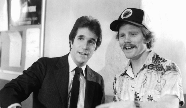 A black and white image depicting Henry Winkler in a suit and in-character during the making of the 1982 film Night Shift talking with director Ron Howard who is wearing a casual shirt and baseball cap.