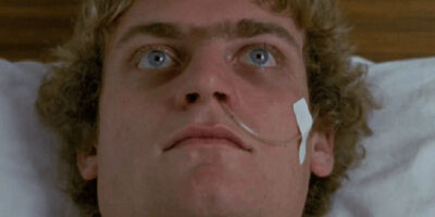 An image from the film Patrick which sees a man in a hospital bed, his eyes wide open, and a nasogastric tube inserted in his nose and secured by white tape on his left cheek.