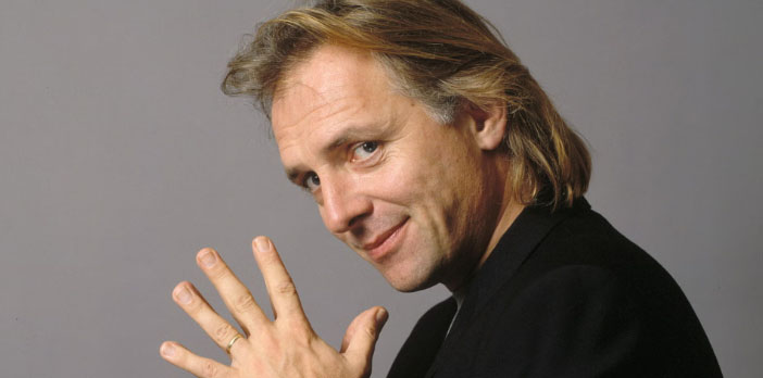 Rik Mayall holds five fingers up to the came and smiles.