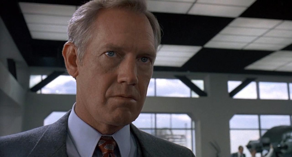 Ronny Cox, his head and shoulders in frame, looking angry in a tailored suit in the film Robocop.