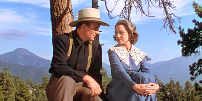 John Wayne and Betty Field sitting on a rock in a picturesque countryside setting in the 1941 film, The Shepherd of the Hills.