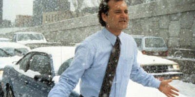Bill Murray wearing a light blue shirt and dark tie standing on a busy road during a snow storm as the traffic has ground to a standstill in the film Groundhog Day.