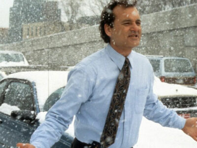 Bill Murray wearing a light blue shirt and dark tie standing on a busy road during a snow storm as the traffic has ground to a standstill in the film Groundhog Day.