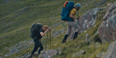 A cinematic still from the film The North, showing two hikers (actors Carles Pulido and Bart Harder) with large backpacks and trekking poles struggling up a steep, rocky, grassy mountain slope in the Scottish Highlands.