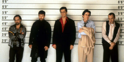 The Usual Suspects iconic police line-up scene featuring Kevin Pollak, Benicio del Toro, Kevin Spacey, Gabriel Byrne, and Stephen Baldwin stood in front of a wall with height measurements on it.