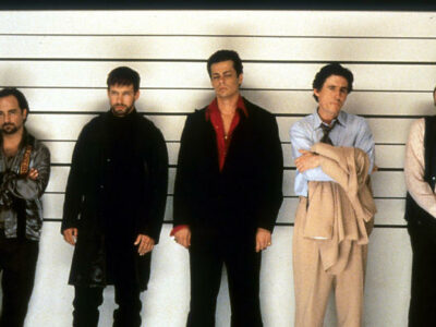 The Usual Suspects iconic police line-up scene featuring Kevin Pollak, Benicio del Toro, Kevin Spacey, Gabriel Byrne, and Stephen Baldwin stood in front of a wall with height measurements on it.