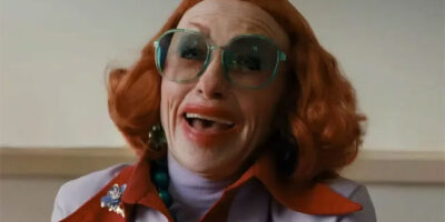 Amy Madigan with fiery red hair, blue-tinged large spectacles and overdone red lipstick in the horror film Weapons.