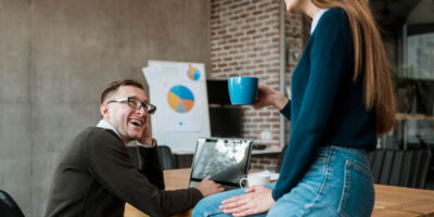 Generic office image with a woman holding a cup of coffee sitting on a desk and chatting with a bespectacled seated man.